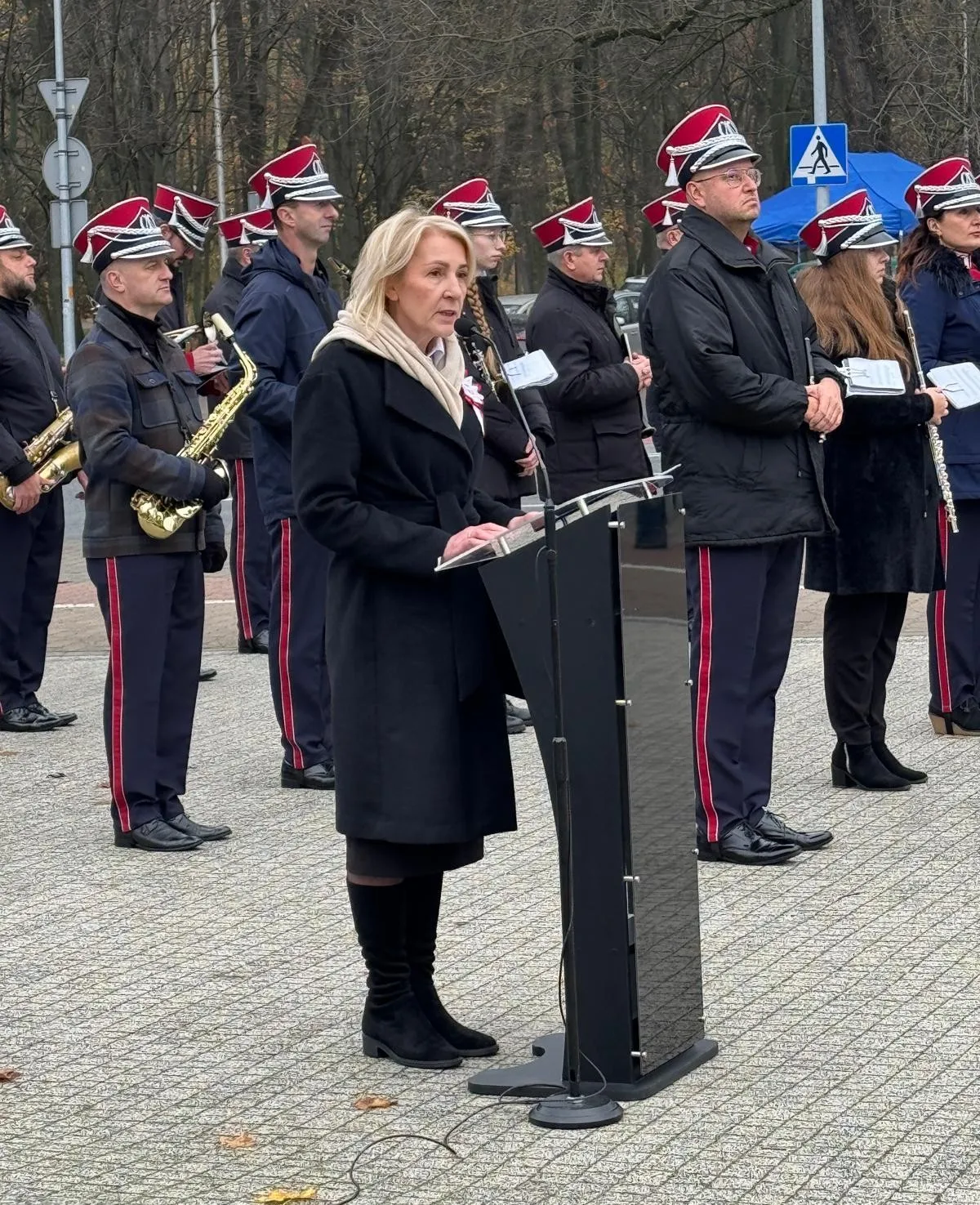 Kobieta w czarnym płaszczu i jasnym szaliku przemawia przy czarnym mównicy na zewnątrz, za nią stoi grupa muzyków w mundurach z czerwonymi czapkami, niektórzy trzymają instrumenty dęte. W tle drzewa bez liści i znak przejścia dla pieszych.